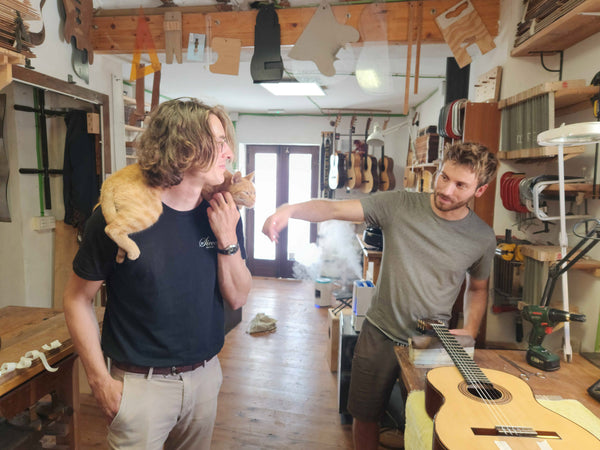 Two men in a guitar shop with one holding a cat and the other playing a guitar.