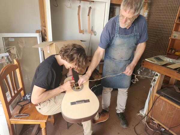 Two people working on a guitar in a workshop setting.