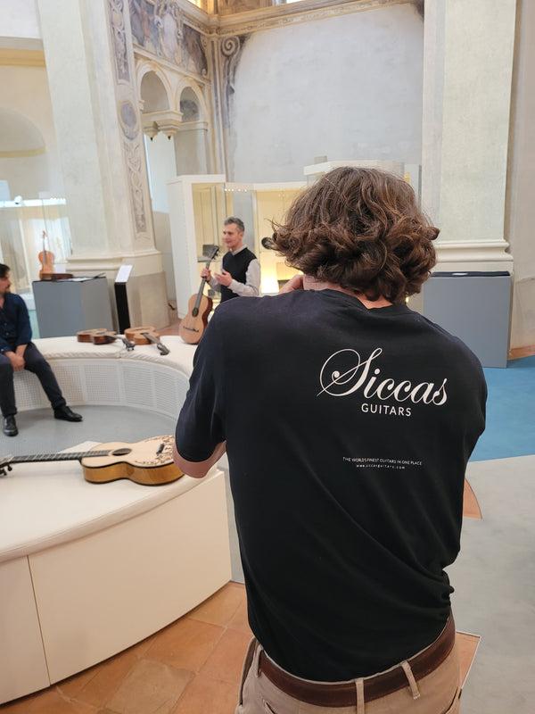 Person wearing a black t-shirt with 'Siccas Guitars' logo, standing in a room with musical instruments.
