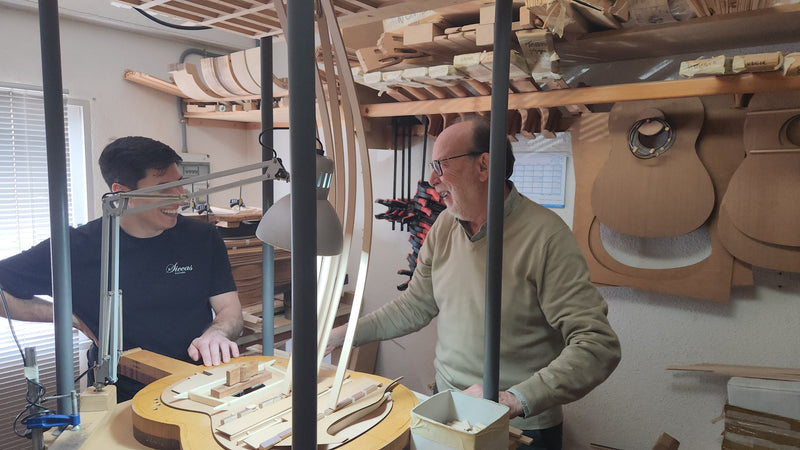 Two men working together in a workshop with musical instruments on shelves.