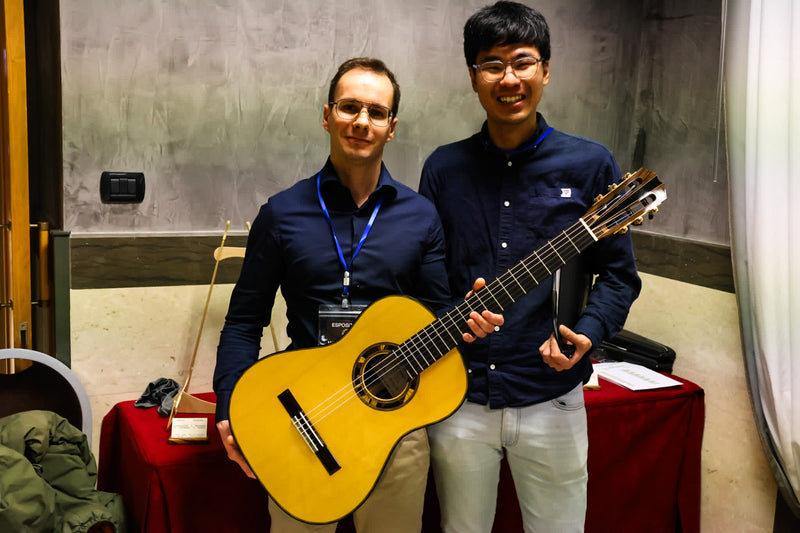 Two men holding a guitar indoors with a neutral background