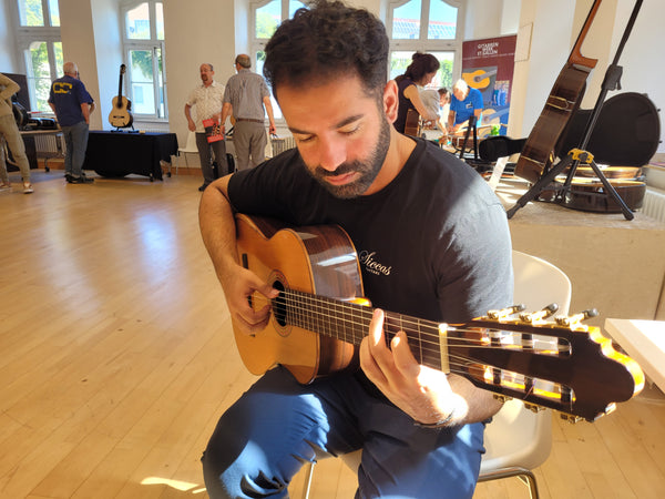 Man playing an acoustic guitar in a room with other people and guitars in the background