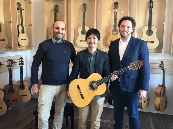 Three men standing in a guitar store with a guitar, surrounded by guitars on display.