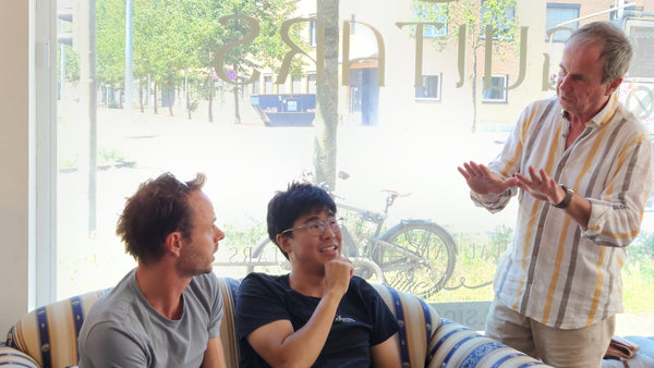 Three men sitting on a couch in a room with large windows, one of them is gesturing.